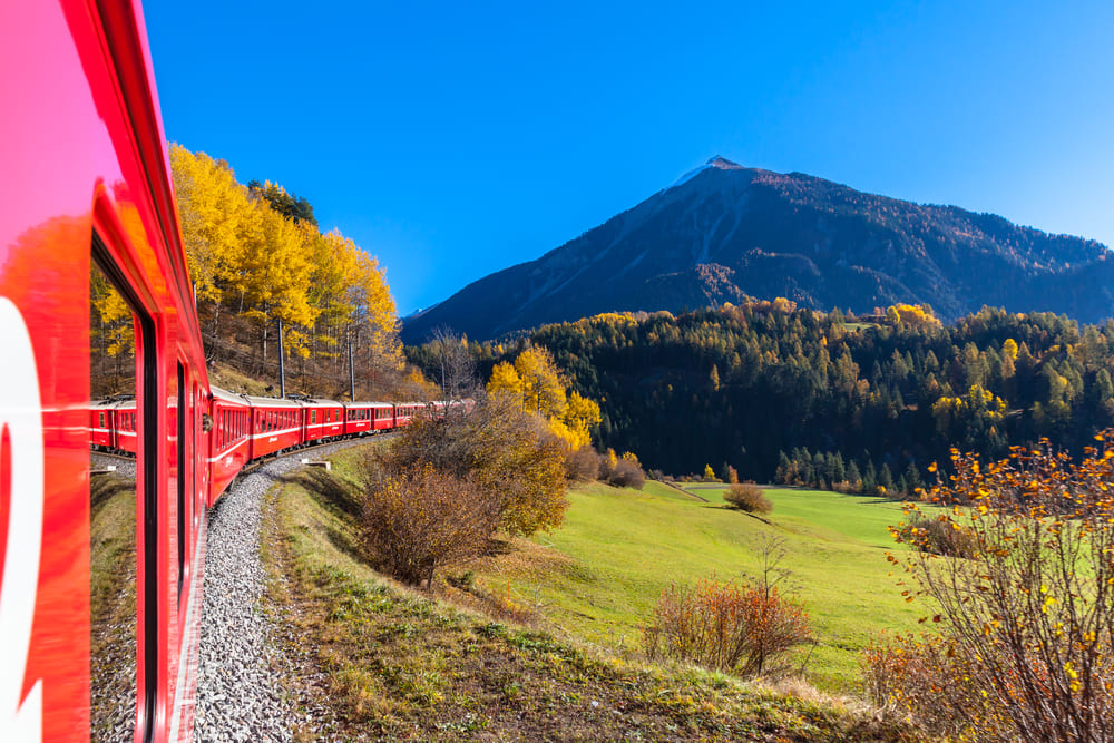 Viaggio in **treno in Europa in autunno** con un treno rosso che attraversa paesaggi montani, circondato da alberi dai colori autunnali e montagne imponenti sotto un cielo blu.