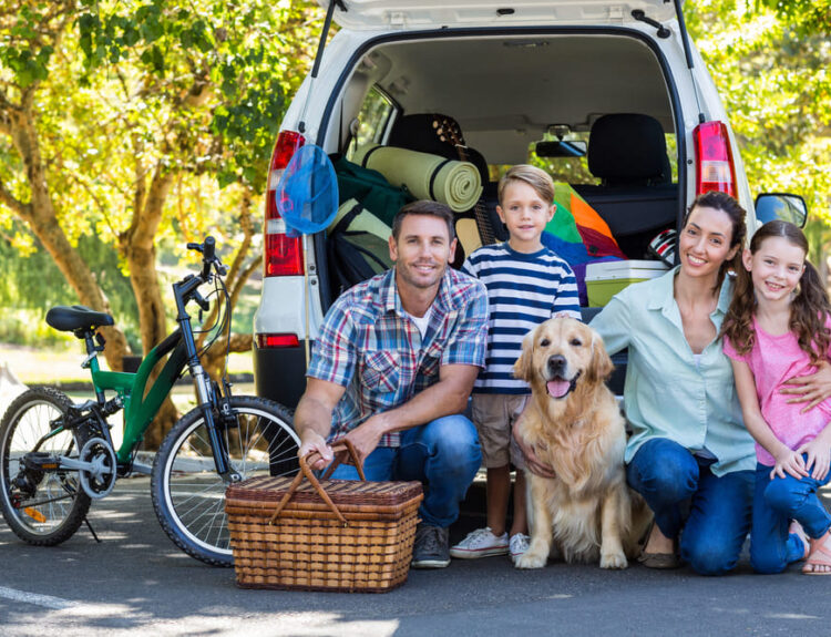 Famiglia sorridente pronta per un viaggio con auto carica, biciclette e un cane. Consigli per viaggiare in famiglia senza stress con i bambini.