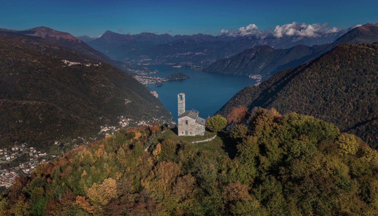 Vista panoramica della Valle d'Intelvi in autunno, con una chiesa isolata circondata da alberi dai colori autunnali e il Lago sullo sfondo tra le montagne.