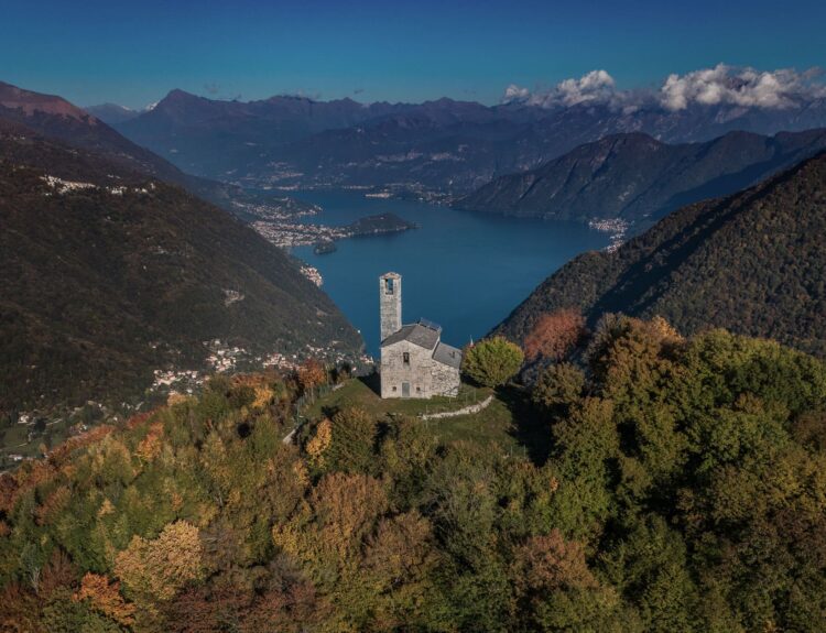 Vista panoramica della Valle d'Intelvi in autunno, con una chiesa isolata circondata da alberi dai colori autunnali e il Lago sullo sfondo tra le montagne.