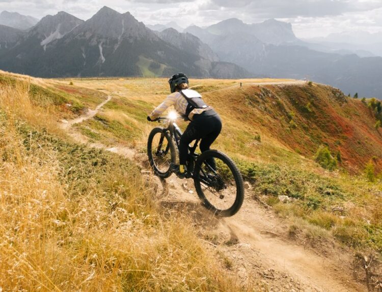 Biker che percorre un sentiero di montagna al Bike Park di Plan de Corones durante l'autunno, circondato da prati e alberi con fogliame dai colori caldi, con montagne sullo sfondo.